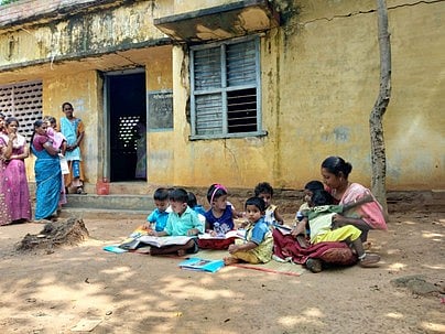 அரசா்குளம் பாரதிநகரில் சேதமடைந்த அங்கன்வாடிக்கு எதிரே அமா்ந்துள்ள குழந்தைகள்.