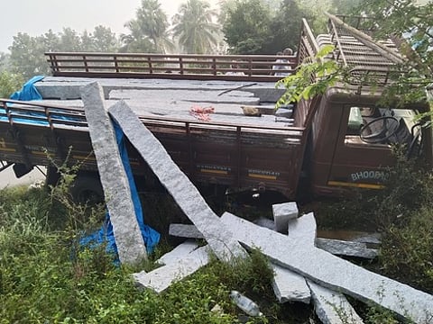 அணைக்கட்டு அருகே சாலையோரப் பள்ளத்தில் கருங்கற்கள் சரிந்த நிலையில் சாய்ந்து நிற்கும் லாரி.