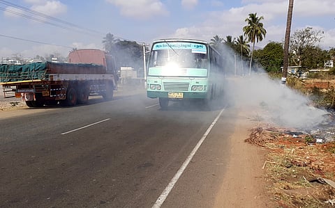 உத்தமபாளையம் தேசிய நெடுஞ்சாலையோரம் வைக்கப்பட்ட தீயால் வெளியேறும் புகையை கடந்து செல்லும் அரசுப் பேருந்து.