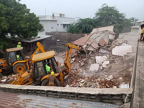 கட்டிட இடிபாடுக்குள் சிக்கிய மூதாட்டியின் பொக்லைன் இயந்திரம் மூலம் உடலை மீட்கும் பணியில் ஈடுபட்ட போலீசார் மற்றும் தீயணைப்பு வீரர்கள்.