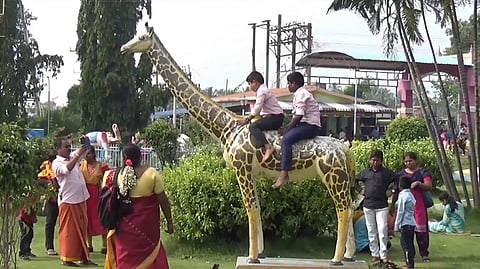 அணைப் பூங்காவில் உள்ள ஒட்டகச்சிவிங்கி சிலை மீது அமா்ந்து விளையாடிய சிறுவா்கள்.