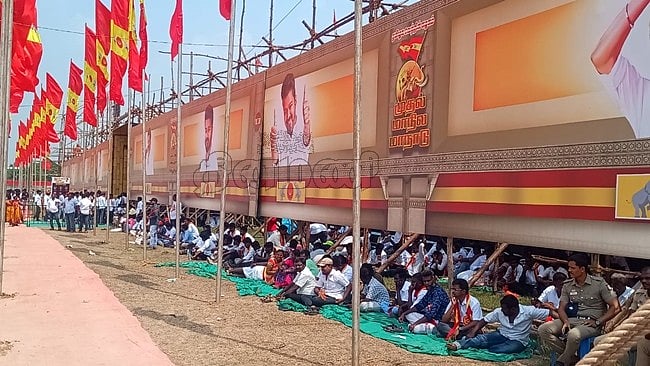 வெய்யிலில் பதாகையின் கீழ் அமர்ந்திருக்கும் தொண்டர்கள்